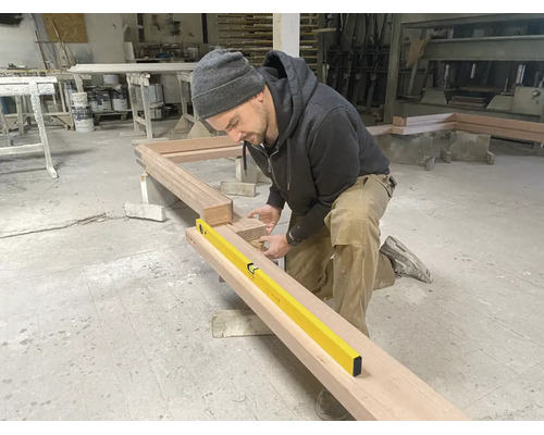 Ein Handwerker in einer Werkstatt prüft mit einer gelben Wasserwaage die Ausrichtung eines Holzrahmens.