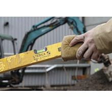 Eine Hand reinigt eine Wasserwaage mit Stabila Logo mit einem Schwamm auf einer Baustelle vor einem Bagger.