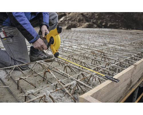 Ouvrier du bâtiment sur armature en acier mesurant avec un mètre ruban jaune la distance par rapport à un coffrage en bois sur un chantier.