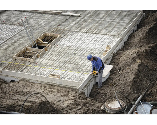Ouvrier du bâtiment sur un chantier mesurant une fondation en béton avec une armature en acier à l'aide d'un mètre ruban jaune.