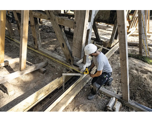 Un ouvrier avec un casque blanc mesure une structure en poutres de bois avec un mètre ruban jaune sur un chantier.
