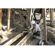 Bauarbeiter mit weißem Schutzhelm misst eine Holzkonstruktion mit einem gelben Maßband auf einer Baustelle.