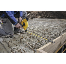 Ouvrier du bâtiment sur armature en acier mesurant avec un mètre ruban jaune la distance par rapport à un coffrage en bois sur un chantier.