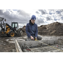 Un ouvrier avec casque de protection mesure une fondation en béton armé à l'aide d'un mètre ruban sur un chantier avec une pelleteuse.