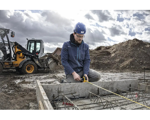Un ouvrier avec casque de protection mesure une fondation en béton armé à l'aide d'un mètre ruban sur un chantier avec une pelleteuse.