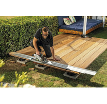 Un artisan coupe des lames de terrasse en bois avec une scie circulaire et un rail de guidage dans le jardin.