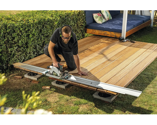 Un artisan coupe des lames de terrasse en bois avec une scie circulaire et un rail de guidage dans le jardin.
