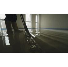 Une personne nivelle un composé de ragréage liquide fraîchement coulé dans une pièce lumineuse avec un long outil de lissage.