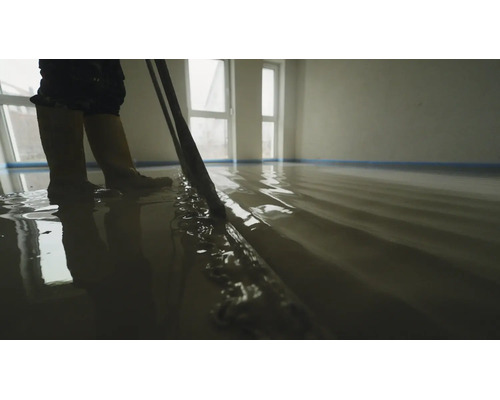 Une personne nivelle un composé de ragréage liquide fraîchement coulé dans une pièce lumineuse avec un long outil de lissage.