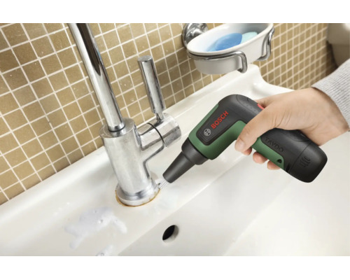 Une main utilise la brosse de nettoyage sans fil Bosch UniversalBrush sur la base d'un robinet de lavabo chromé.