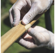 Hände mit Handschuhen tragen Holzspachtelmasse zur Reparatur auf ein Holzstück auf.
