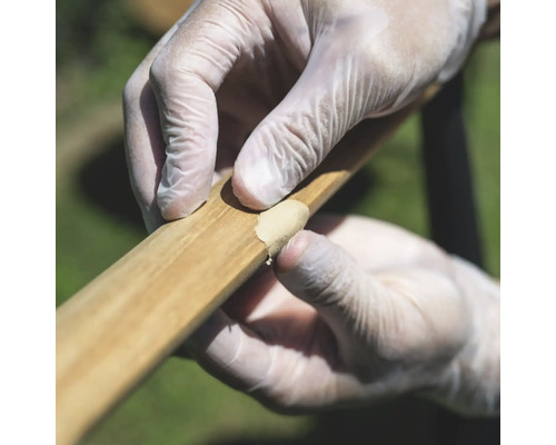 Hände mit Handschuhen tragen Holzspachtelmasse zur Reparatur auf ein Holzstück auf.