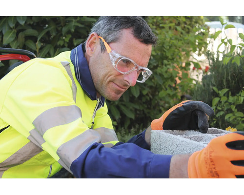Homme en veste haute visibilité avec lunettes de protection et gants de travail travaillant sur un bloc de béton.