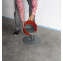 Une personne verse un composé de nivellement gris d'un seau rouge sur un sol en béton pour niveler le sol.