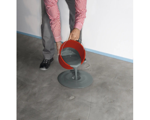 Une personne verse un composé de nivellement gris d'un seau rouge sur un sol en béton pour niveler le sol.