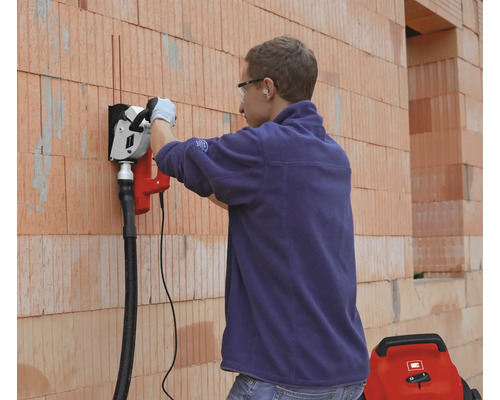 Une personne fraise des rainures dans un mur en briques avec une rainureuse à béton raccordée à un aspirateur.