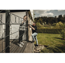Un homme installe une clôture décorative noire sur une terrasse avec une perceuse sans fil, une femme se tient à ses côtés dans un jardin.