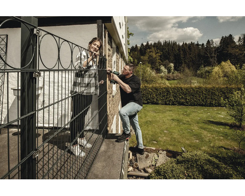 Un homme installe une clôture décorative noire sur une terrasse avec une perceuse sans fil, une femme se tient à ses côtés dans un jardin.