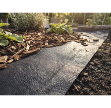 Toile de paillage noire dans un massif de jardin, partiellement recouverte d écorce de bois, à côté de terre et de petites plantes.