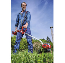 Faux sans fil Einhell en action : un homme fauche de l'herbe haute dans un jardin en portant un équipement de protection et une sangle.
