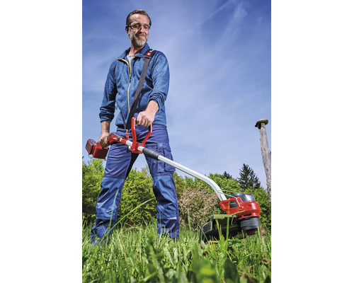 Faux sans fil Einhell en action : un homme fauche de l'herbe haute dans un jardin en portant un équipement de protection et une sangle.