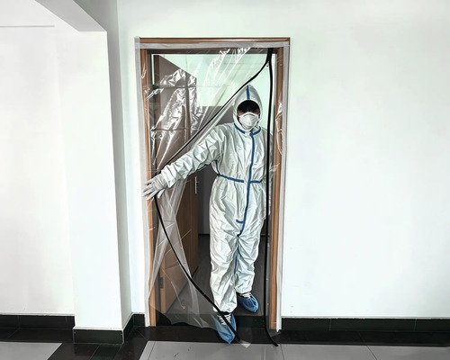 Porte de protection contre la poussière transparente avec fermeture éclair incurvée fixée sur un cadre de porte, personne en tenue de protection.
