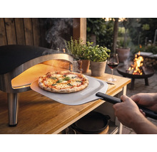 Frisch gebackene Pizza wird mit einem Pizzaheber mit Tenneker Logo aus einem tragbaren Pizzaofen auf einem Holztisch im Garten entnommen.