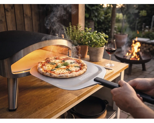 Frisch gebackene Pizza wird mit einem Pizzaheber mit Tenneker Logo aus einem tragbaren Pizzaofen auf einem Holztisch im Garten entnommen.