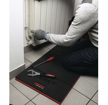 Personne à genoux sur un tapis de protection Virax travaillant sur un radiateur avec un tournevis. Des outils sont posés sur le tapis.