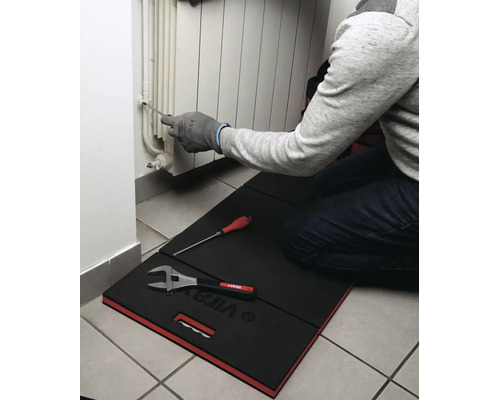 Personne à genoux sur un tapis de protection Virax travaillant sur un radiateur avec un tournevis. Des outils sont posés sur le tapis.