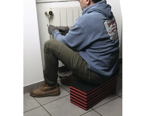 Homme assis sur un repose-genoux modulaire à rayures rouges et noires travaillant sur un radiateur.
