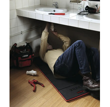 Plombier allongé sur un tapis de genoux Virax sous un lavabo pour réparer le siphon. Sac à outils et coupe-tube sont posés au sol.