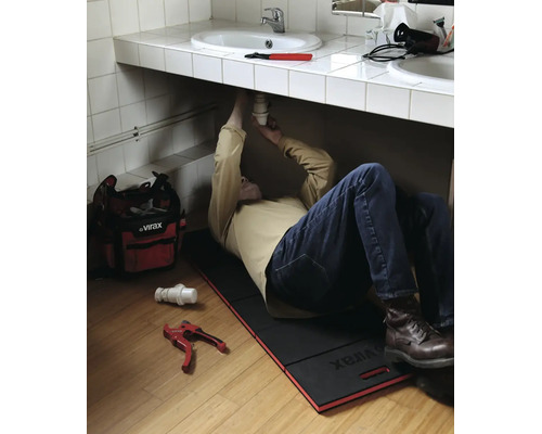 Plombier allongé sur un tapis de genoux Virax sous un lavabo pour réparer le siphon. Sac à outils et coupe-tube sont posés au sol.