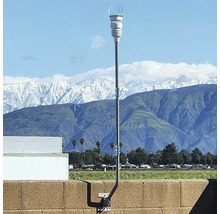 Station météo avec capteur blanc sur un mât métallique fixé à un mur, devant un paysage de montagne lumineux aux sommets enneigés.