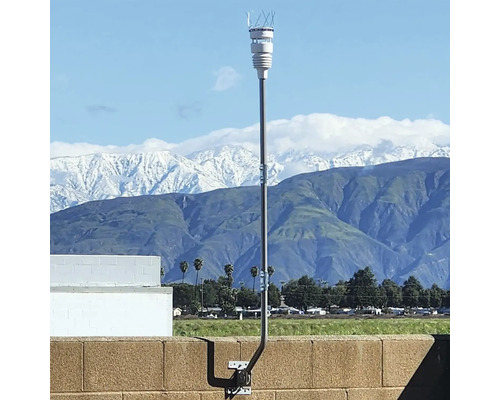 Station météo avec capteur blanc sur un mât métallique fixé à un mur, devant un paysage de montagne lumineux aux sommets enneigés.