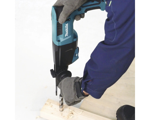 Une personne portant des gants de protection utilise un perforateur Makita pour percer un morceau de bois. Logo Makita visible.