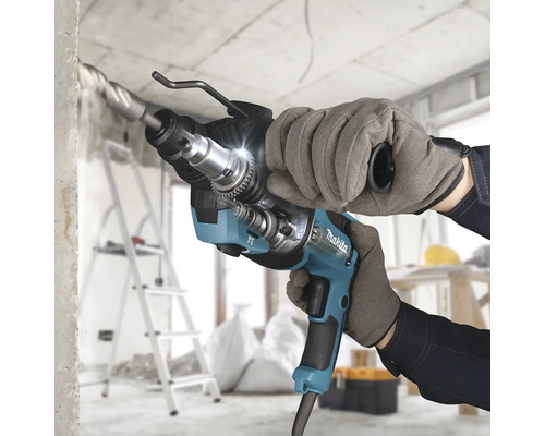 Marteau perforateur Makita perçant du béton sur un chantier lumineux, tenu par une personne avec des gants de travail.