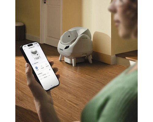 Person steuert eine selbstreinigende Cat Link Katzentoilette per Applikation auf einem Smartphone in einem hellen Raum mit Holzboden.