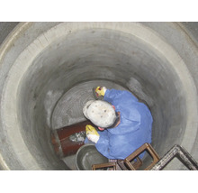 Un ouvrier en combinaison de protection bleue scelle le fond d'un regard en béton avec du mortier à côté d'un tuyau.