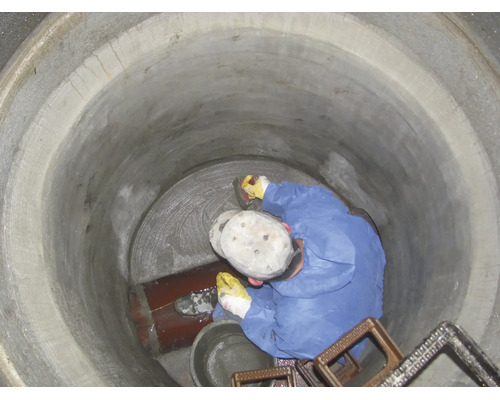 Un ouvrier en combinaison de protection bleue scelle le fond d'un regard en béton avec du mortier à côté d'un tuyau.