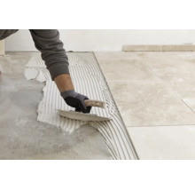 Une personne applique de la colle à carrelage sur le sol avec une spatule crantée pour poser des carreaux de sol aspect marbre.