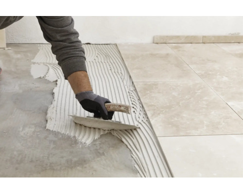 Une personne applique de la colle à carrelage sur le sol avec une spatule crantée pour poser des carreaux de sol aspect marbre.