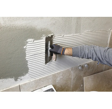 Une main gantée applique de la colle à carrelage sur un mur crépi à l'aide d'une truelle dentelée.
