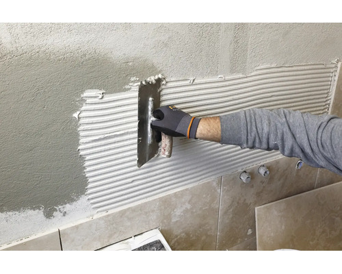 Une main gantée applique de la colle à carrelage sur un mur crépi à l'aide d'une truelle dentelée.