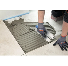 Application de colle à carrelage sur le sol à l'aide d'une spatule crantée pour la pose de carreaux de sol.