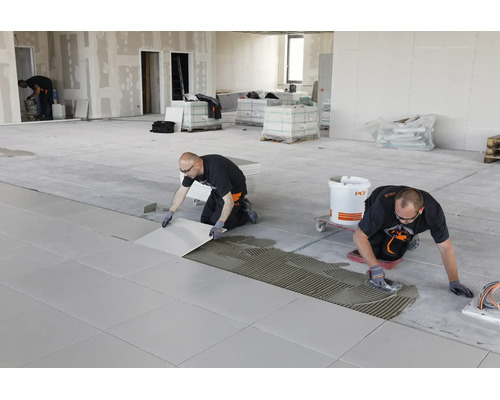 Handwerker verlegen großformatige Bodenfliesen in einem hellen, weitläufigen Raum unter Verwendung von Fliesenkleber und einer Zahnkelle.
