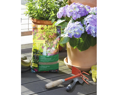 Sac d'engrais spécial jardin Manna sur une terrasse en bois à côté d'un hortensia en pot, d'outils de jardin et de gants.