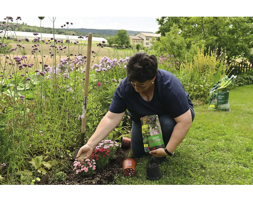 Femme fertilisant un parterre de fleurs avec de l'engrais, accompagnée d'une bêche, de pots et d'un sac de terreau.