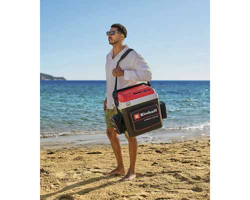 Un homme porte un sac isotherme Einhell avec bandoulière et poches latérales sur une plage de sable lumineuse au bord de la mer. Einhell Logo.