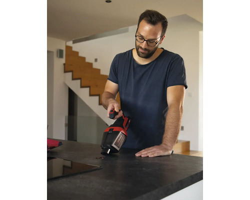 Un homme nettoie des grains de café sur un plan de travail de cuisine à l aide d un aspirateur à main Einhell dans un intérieur moderne.